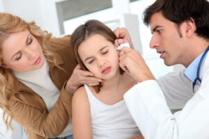 Doctor examining a young girl's ear with an otoscope while her concerned mother supports her, checking for ear infection or hearing issues.