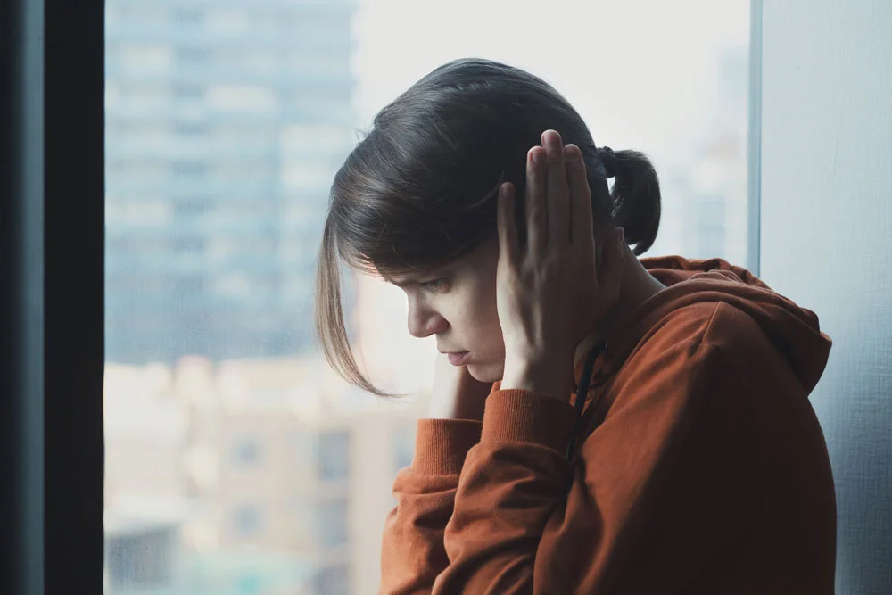 Young brown haired woman sitting next to window holding her hands over her ears