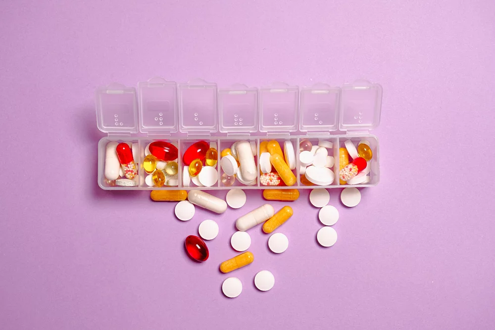 Medicine organiser box with various types of pills spilling out against muted purple background