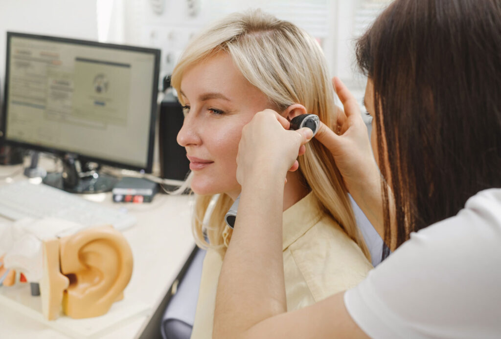 a woman sitting and another woman doing hearing test using otoscopy.